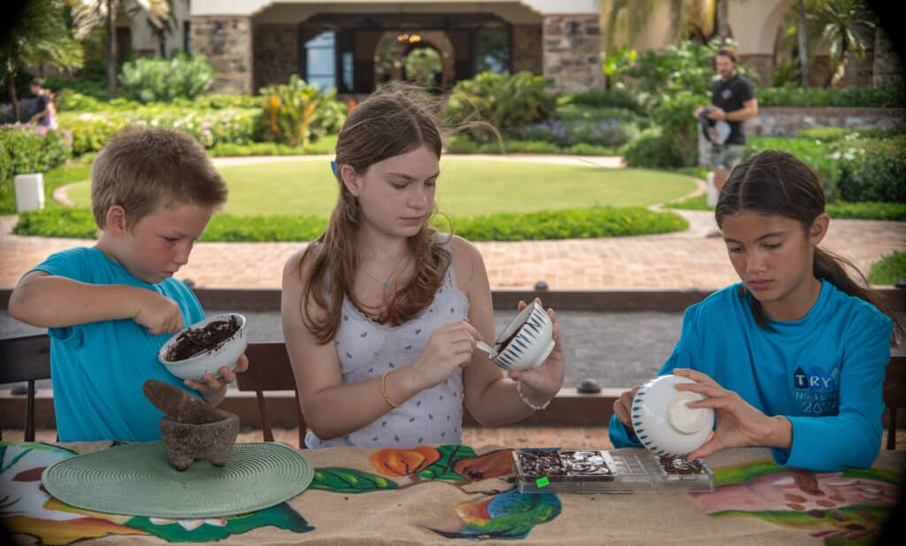 Three children sit at a table outdoors, focused on crafting their own chocolate bars. Each works with bowls and molds to make custom chocolate, surrounded by greenery and a villa in the background.