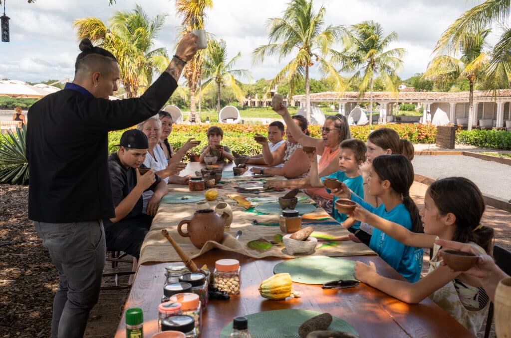 A group of people, including children and adults, sit around a long outdoor table raising cups in a toast, while enjoying chocolate personalizado in a resort-like setting with palm trees in the background.