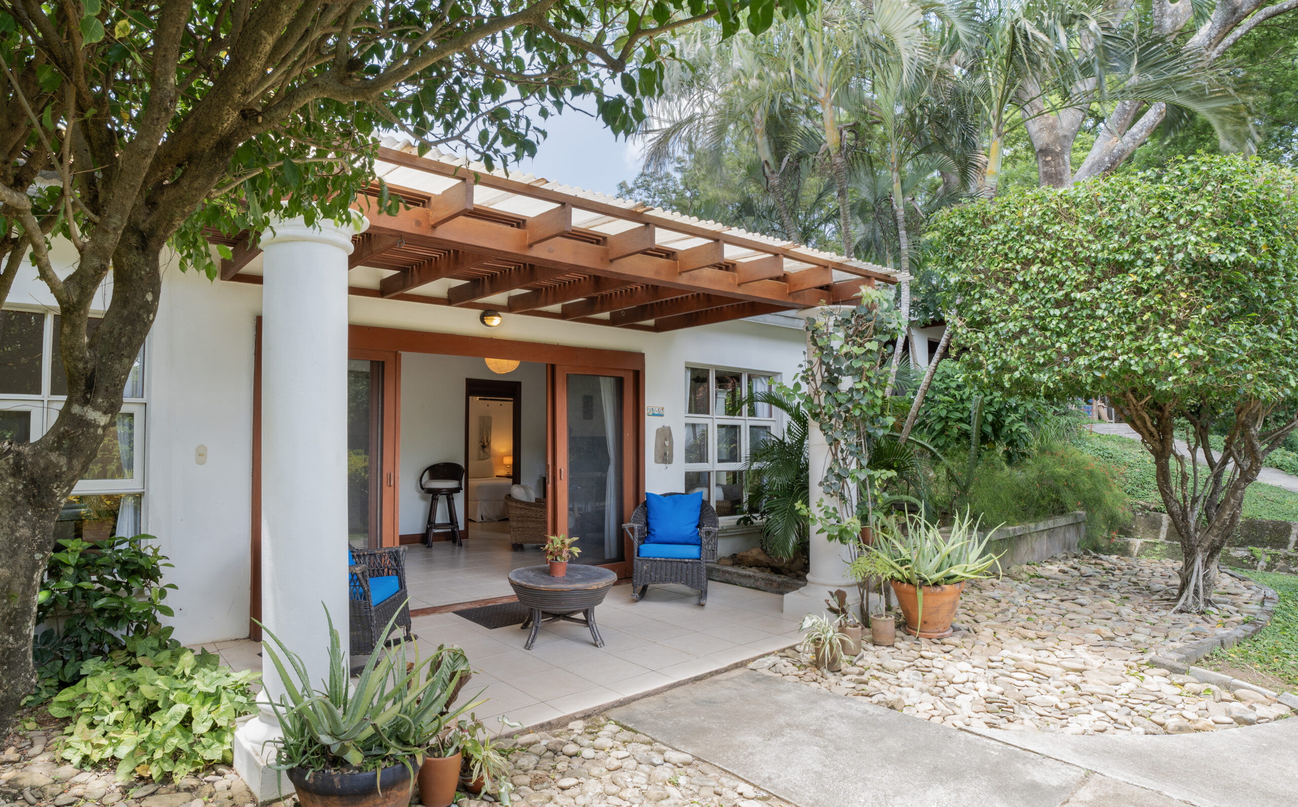 http://A%20cozy%20house%20with%20a%20covered%20front%20porch,%20white%20columns,%20and%20wooden%20beams,%20surrounded%20by%20lush%20greenery,%20potted%20plants,%20and%20a%20stone%20path%20leading%20to%20the%20entrance.%20Blue%20cushions%20decorate%20the%20porch%20seating%20area.