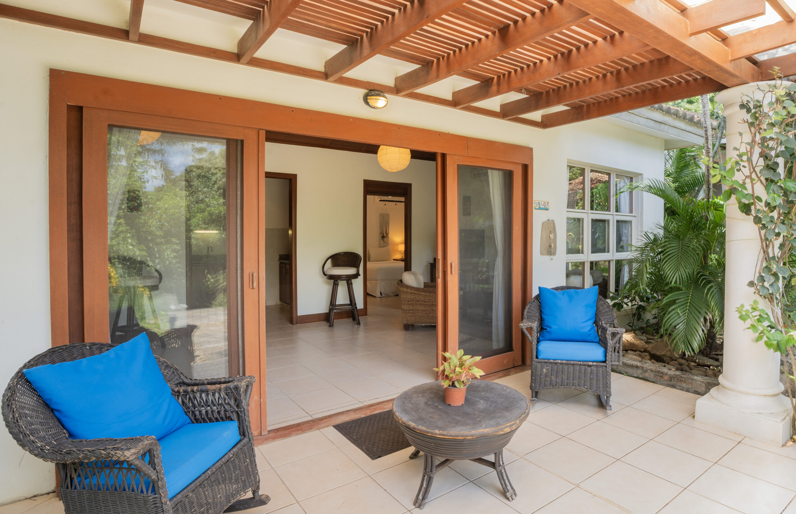 http://A%20covered%20patio%20with%20two%20wicker%20chairs%20with%20blue%20cushions,%20a%20small%20round%20table%20with%20a%20potted%20plant,%20and%20sliding%20glass%20doors%20leading%20to%20a%20bright%20interior%20space.%20Green%20plants%20and%20trees%20surround%20the%20area.