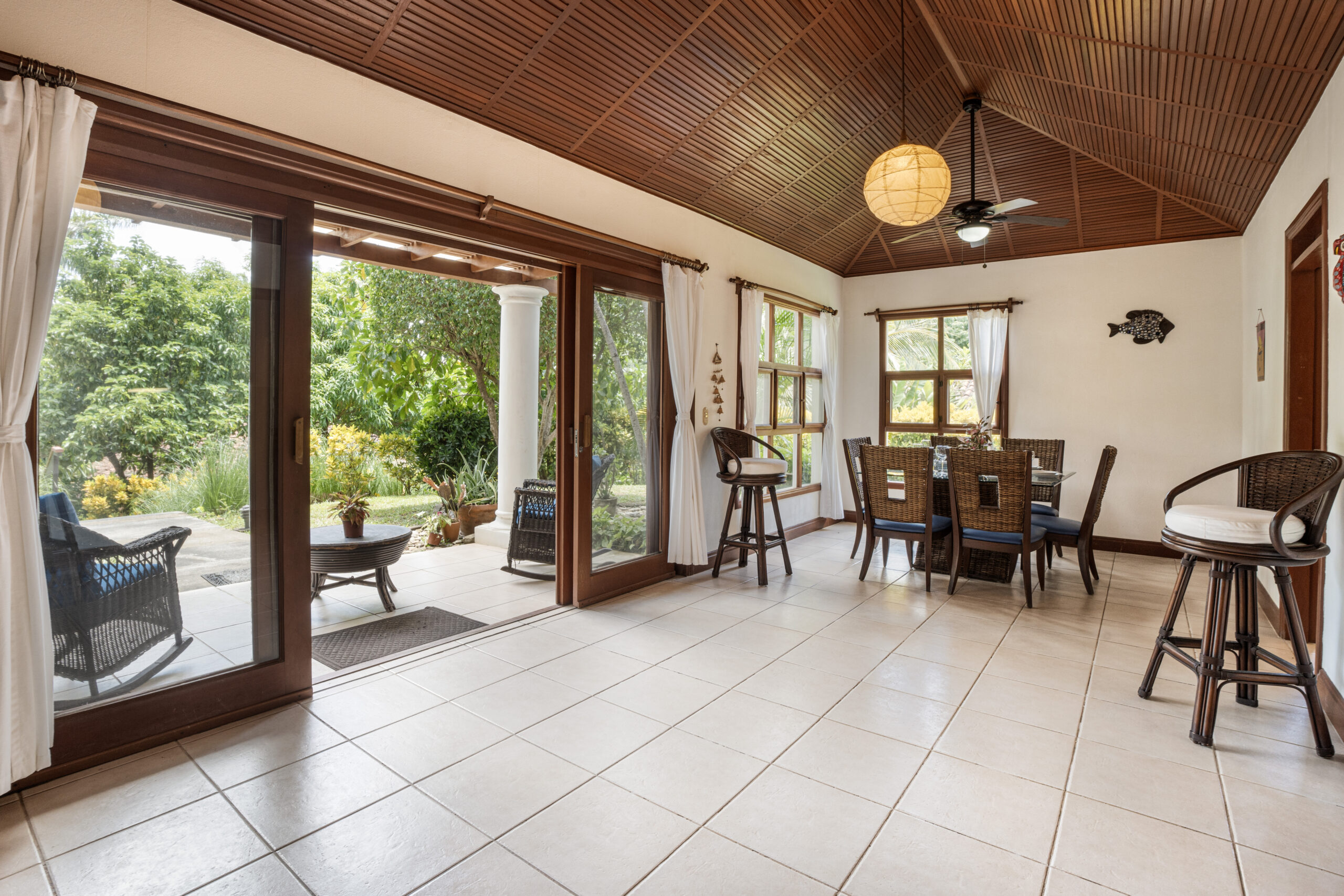 http://Bright%20sunroom%20with%20tiled%20floors,%20wooden%20ceiling,%20large%20glass%20doors%20opening%20to%20a%20garden,%20a%20dining%20table%20with%20chairs,%20bar%20stools,%20and%20natural%20light%20flooding%20the%20space,%20creating%20a%20cozy%20indoor-outdoor%20atmosphere.
