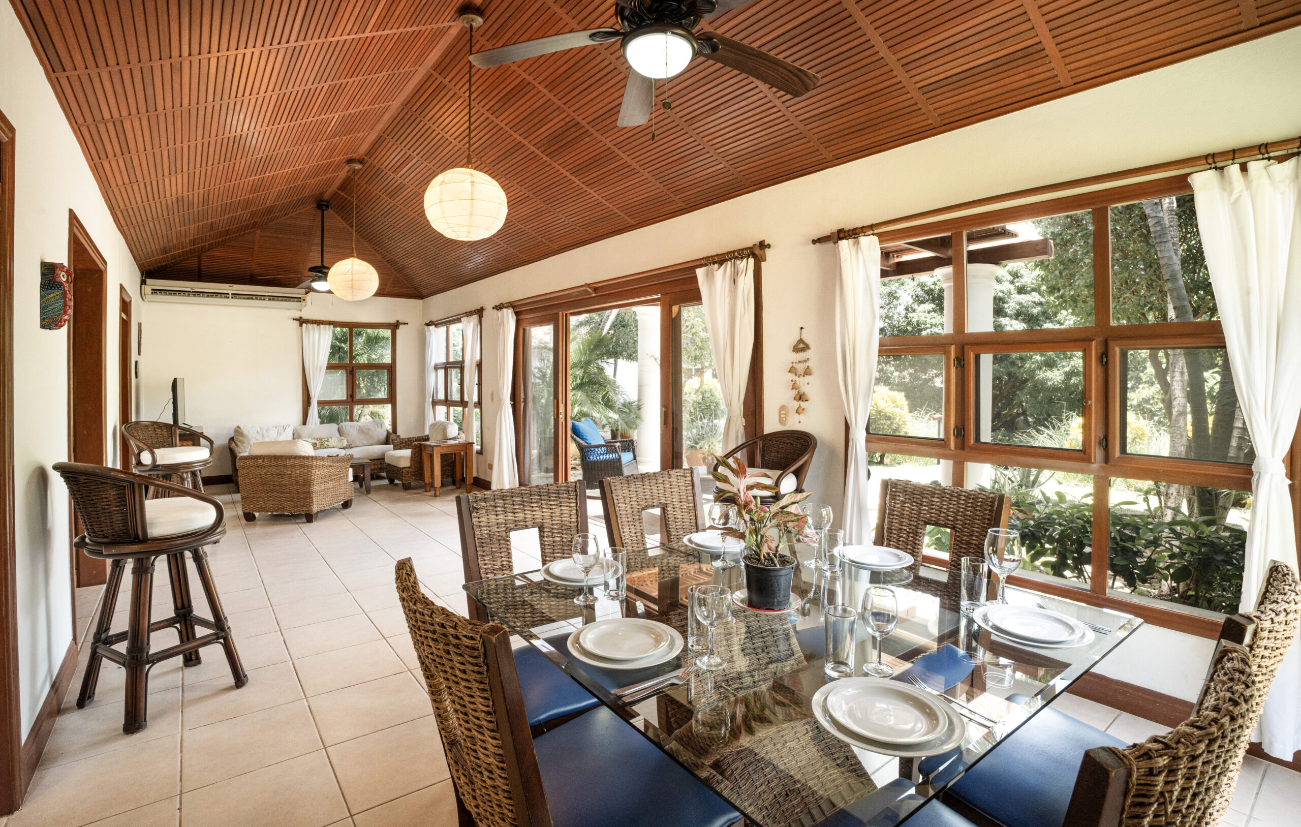 http://A%20bright,%20airy%20dining%20and%20living%20area%20with%20wicker%20chairs,%20a%20glass%20dining%20table%20set%20for%20six,%20large%20windows,%20white%20curtains,%20tiled%20floors,%20ceiling%20fans,%20and%20wooden%20ceilings,%20overlooking%20a%20green%20garden%20outside.