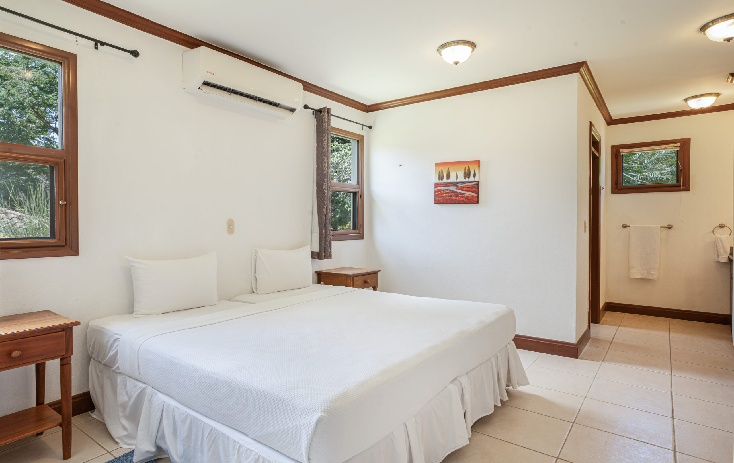 http://A%20bright%20bedroom%20with%20a%20white%20bed,%20two%20wooden%20nightstands,%20windows%20with%20brown%20trim,%20wall%20art%20above%20the%20bed,%20and%20an%20air%20conditioner.%20A%20towel%20hangs%20near%20the%20bathroom%20visible%20in%20the%20background.