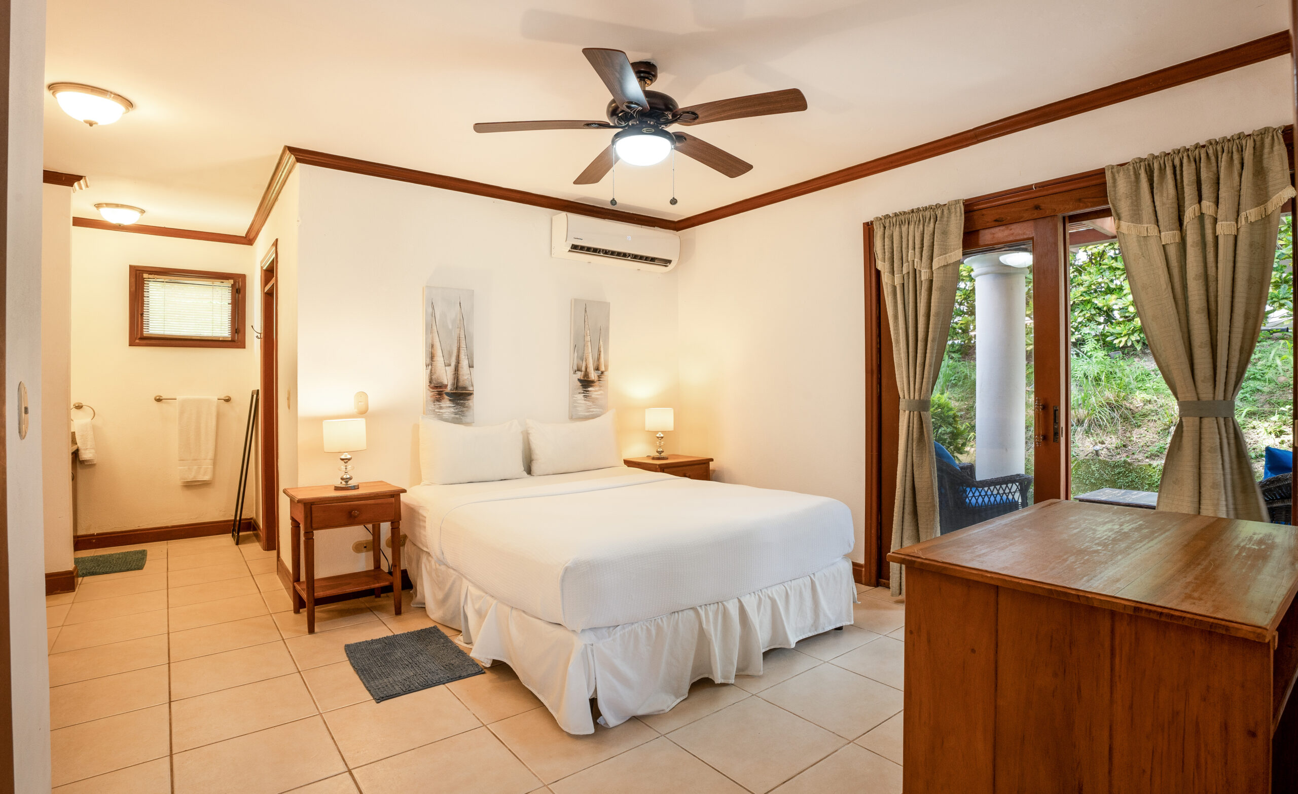 http://A%20cozy%20bedroom%20with%20a%20double%20bed,%20white%20bedding,%20two%20bedside%20tables%20with%20lamps,%20a%20ceiling%20fan,%20wooden%20furniture,%20and%20glass%20doors%20leading%20to%20a%20patio%20with%20greenery%20outside.