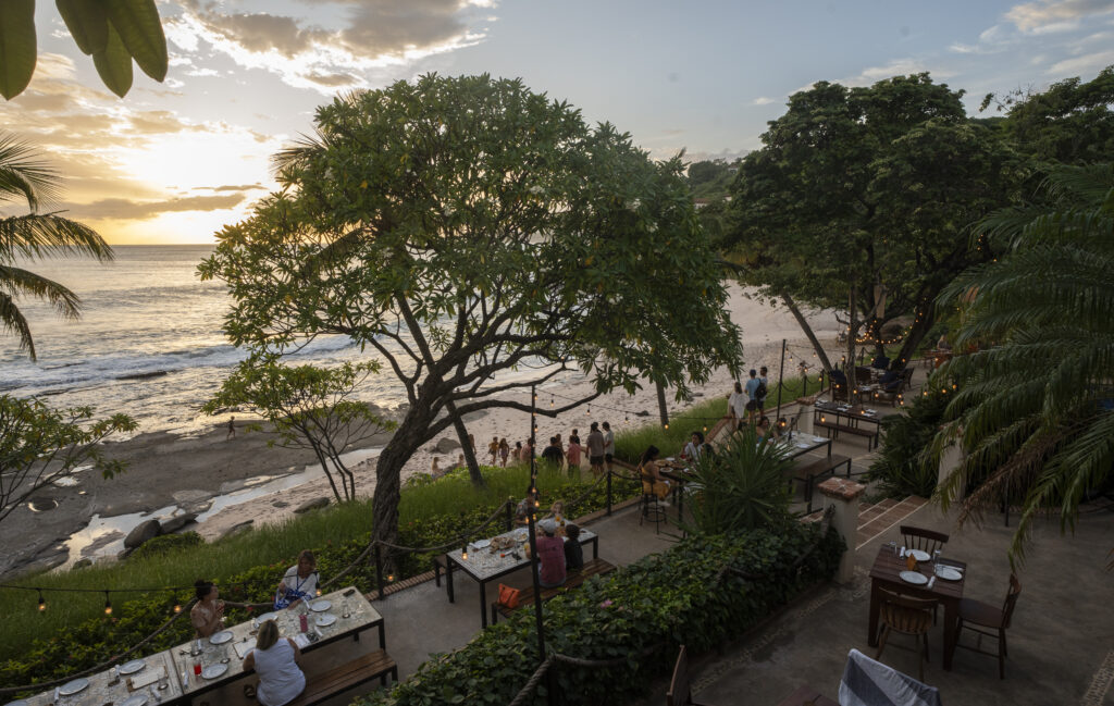 Outdoor restaurant overlooking a beach at sunset, with diners seated at tables among trees and string lights. People are walking along the sandy shore near the ocean, surrounded by lush greenery.