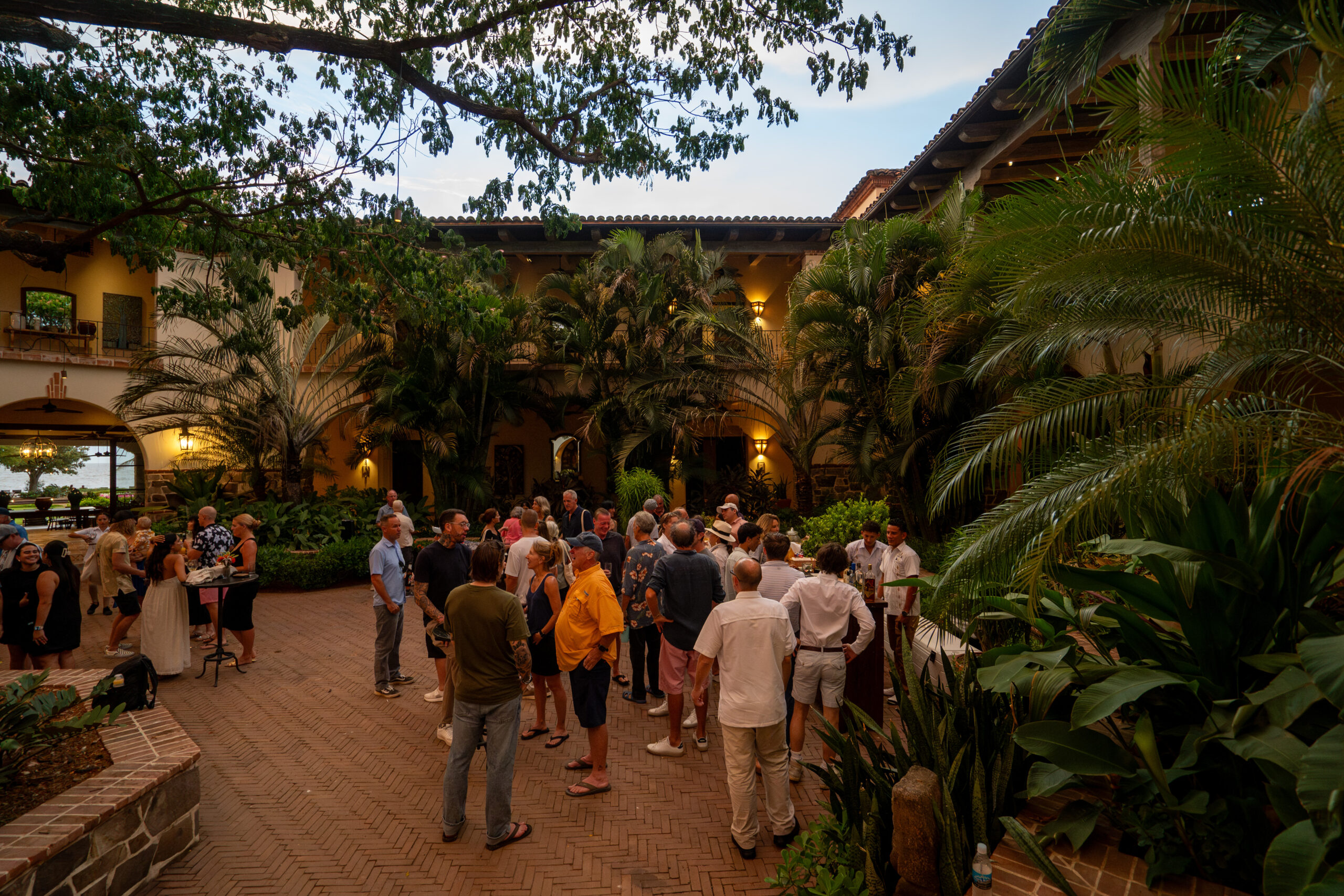 Supporters gather for CREA’s Annual Fundraiser, socializing in a lush, tropical courtyard with brick flooring, surrounded by palm trees and warmly lit buildings in the evening.