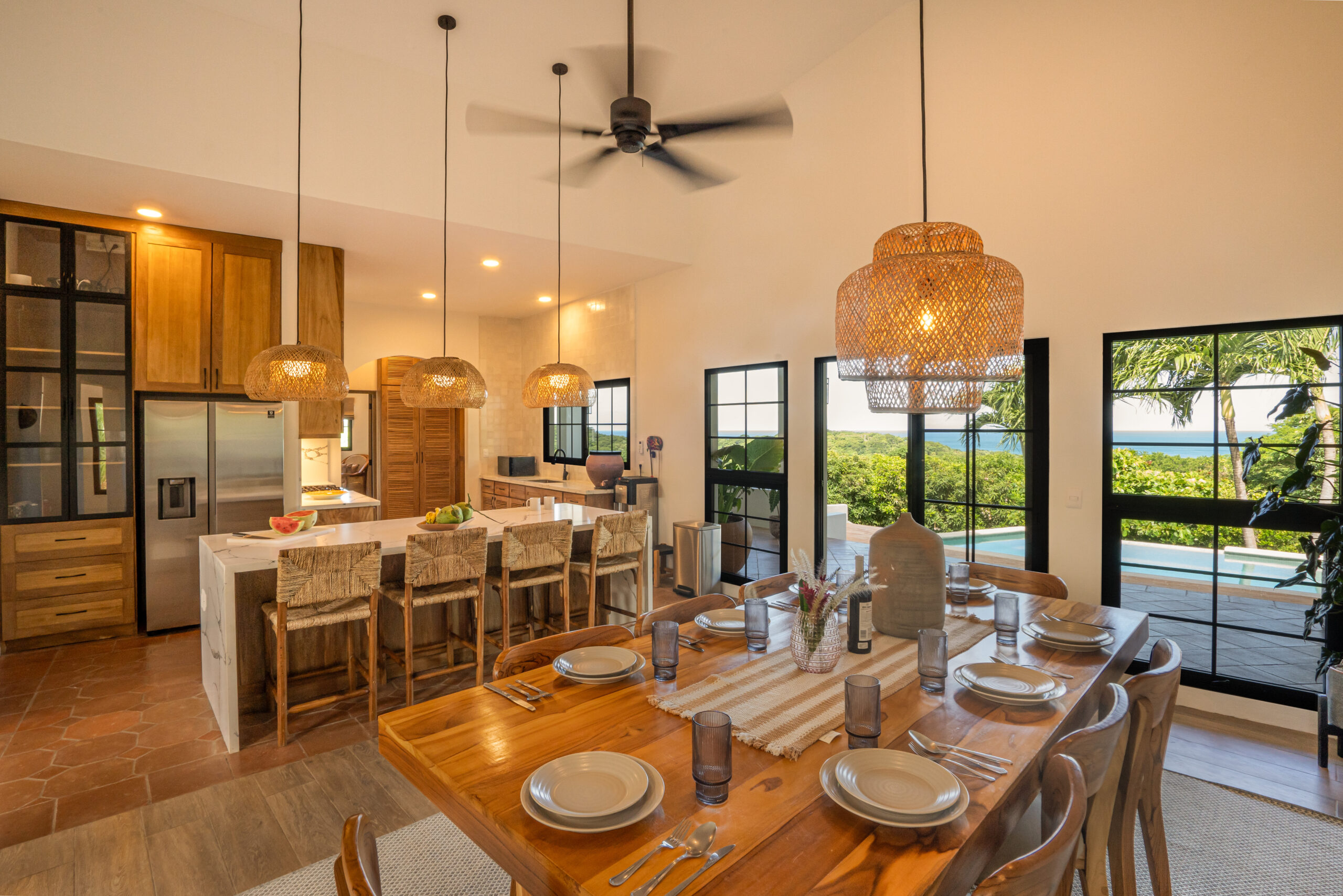 http://A%20spacious,%20modern%20kitchen%20and%20dining%20area%20at%20Casa%20Reposo%20features%20wooden%20cabinets,%20a%20large%20island%20with%20barstools,%20a%20set%20dining%20table,%20woven%20pendant%20lights,%20and%20windows%20framing%20scenic%20views%20of%20palm%20trees%20and%20water.