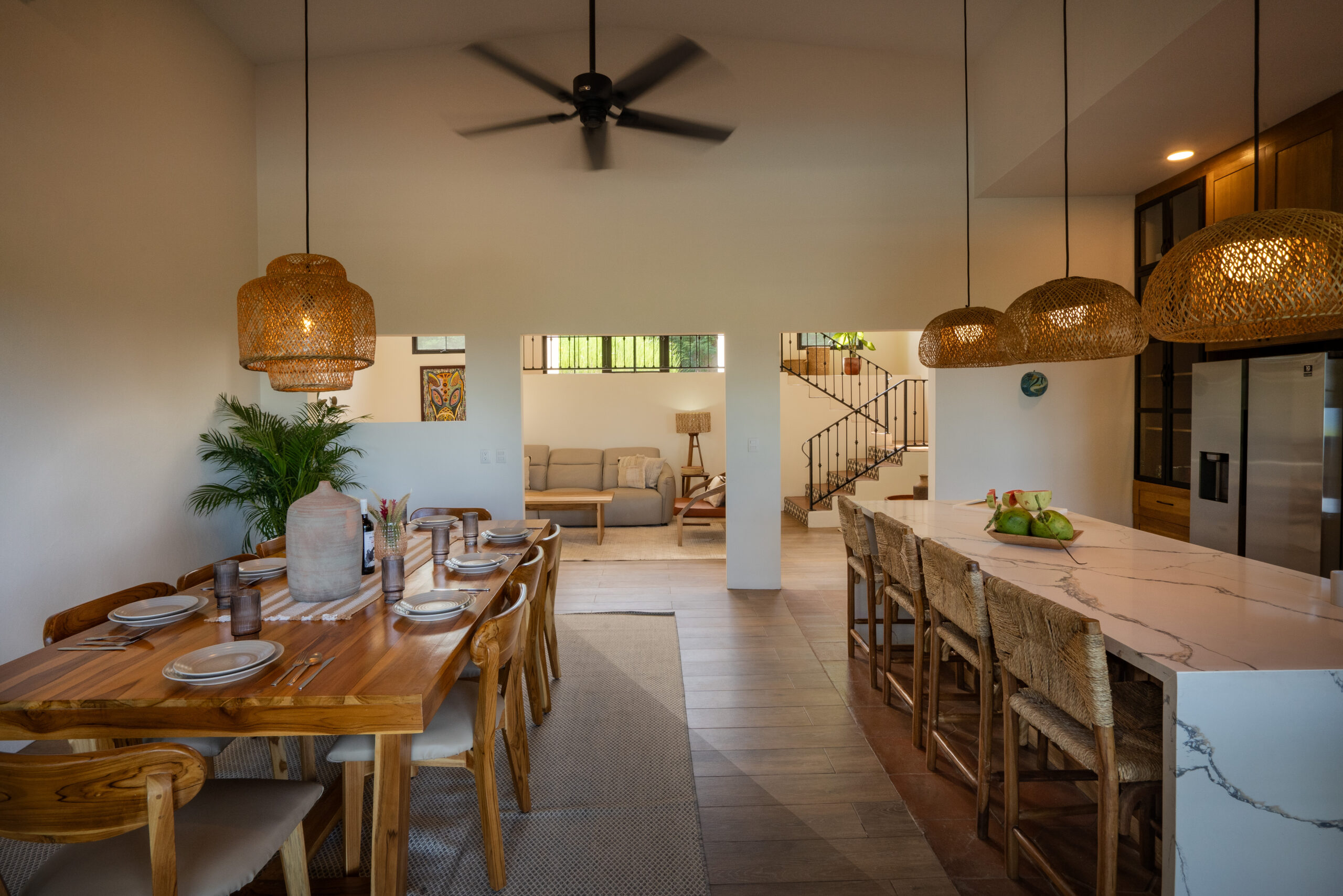 http://Modern%20open-concept%20dining%20and%20kitchen%20area%20in%20Casa%20Reposo,%20featuring%20a%20wooden%20table%20set%20for%20eight,%20marble%20island%20with%20barstools,%20wicker%20pendant%20lights,%20ceiling%20fan,%20and%20a%20view%20into%20a%20cozy%20living%20room%20with%20a%20staircase.