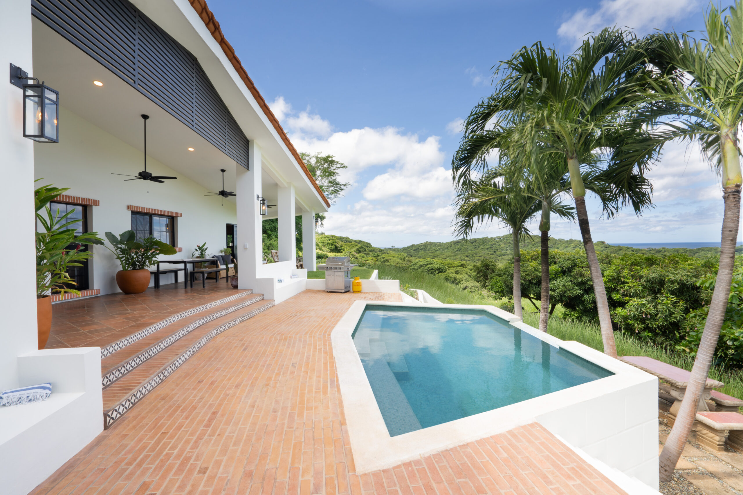 http://A%20modern%20patio%20at%20Casa%20Reposo%20with%20terracotta%20tiles,%20potted%20plants,%20and%20ceiling%20fans%20overlooks%20a%20small%20rectangular%20pool%20and%20lush%20green%20landscape%20under%20a%20blue%20sky%20with%20scattered%20clouds.