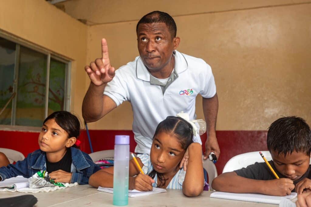 Un profesor hace gestos mientras da instrucciones a tres jóvenes alumnos, que están escribiendo en sus cuadernos en una mesa del aula durante una clase de CREA. Una de las alumnas parece pensativa, mientras que los demás se concentran en su trabajo, lo que refleja la dedicación que contribuye a las iniciativas de la Recaudación Anual.