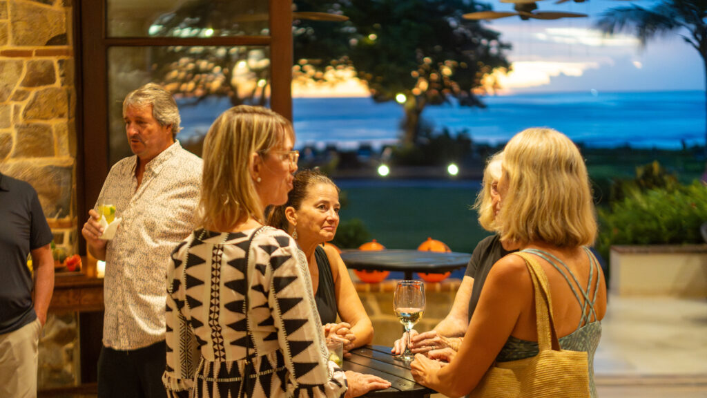 A group of people stand and talk indoors near a window at sunset at Rancho Santana, with a view of the ocean and trees outside. Two women hold wine glasses, creating a relaxed and social vibe during the Green Season.