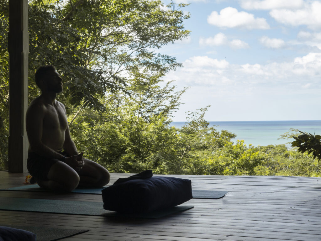 A man sits cross-legged on a yoga mat in a shaded wooden pavilion at Rancho Santana, overlooking lush greenery and a distant ocean view under the partly cloudy sky of Green Season.