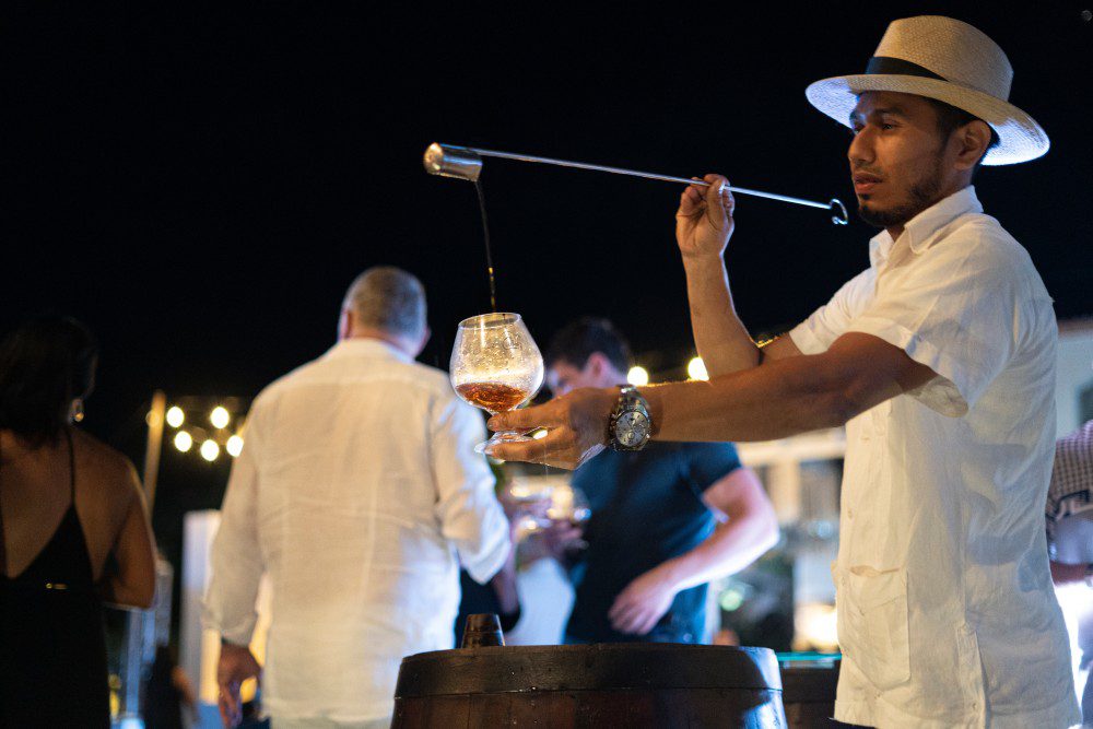 A man in a white shirt and hat pours a drink with a long-handled ladle into a glass at an outdoor Rancho Santana nighttime event, with blurred people and string lights glowing in the Green Season background.
