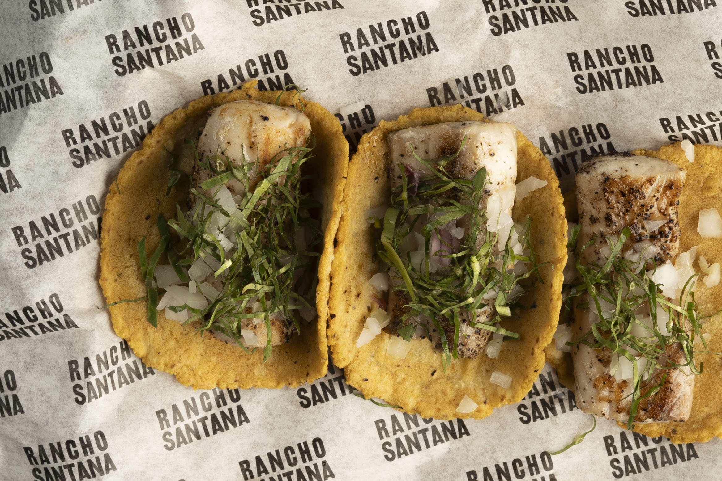 Three fish tacos topped with chopped onions and fresh cilantro are arranged on branded Rancho Santana paper, perfect for savoring during Green Season.