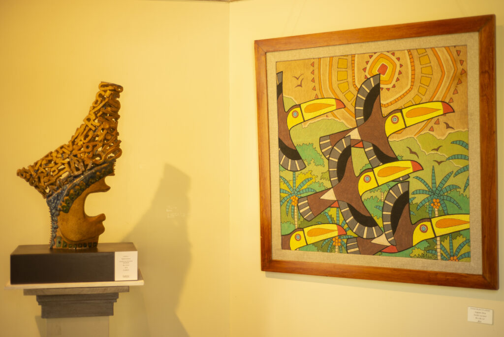 A decorative sculpture on a pedestal stands beside a colorful framed painting of toucans amidst tropical plants on a yellow wall in an art gallery.