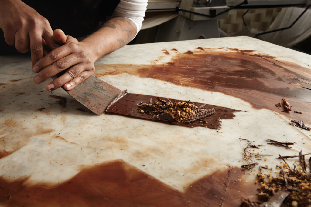 A person uses a large metal scraper to spread and shape melted chocolate with crushed nuts on a marble slab, crafting their own custom chocolate bar as chocolate is smeared across the surface.