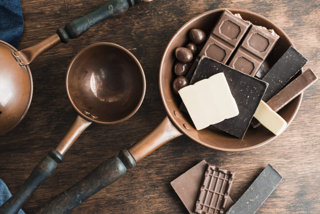 Assorted chocolate bars and pieces, including dark, milk, and white chocolate, are arranged in copper bowls on a rustic wooden surface—perfect inspiration to craft your own chocolate bar with custom flavors. Large wooden-handled ladles sit nearby.