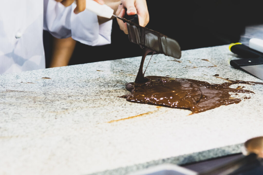 A person in a white coat spreads melted chocolate on a stone countertop using a spatula, likely tempering it to craft your own chocolate bar during the custom chocolate confectionery process.