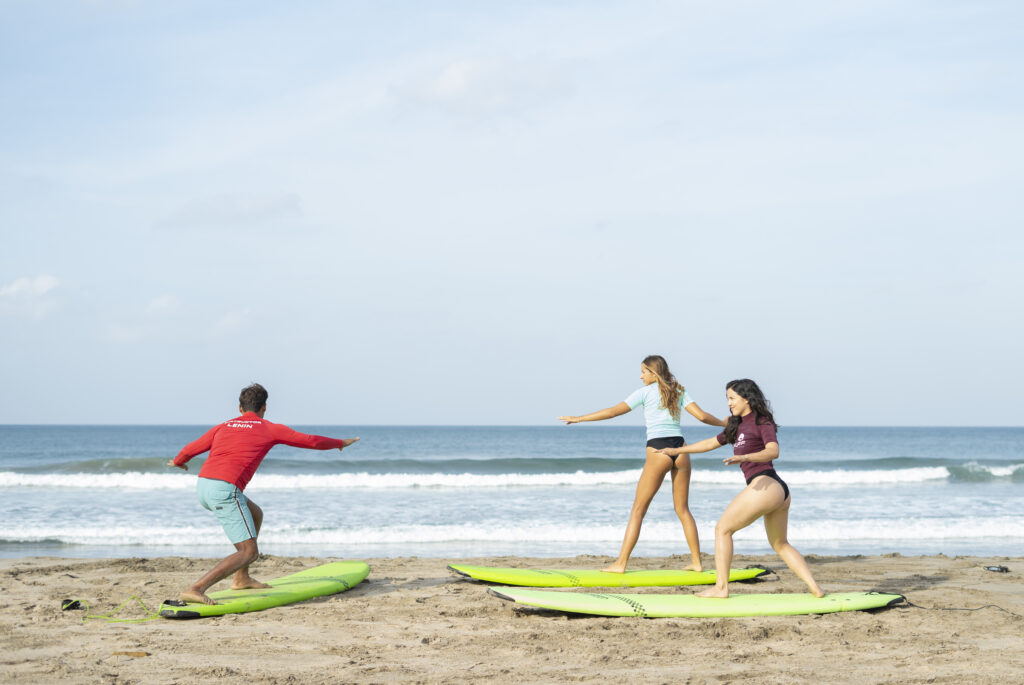 On a sandy Rancho Santana beach, three people practice surfing moves on green surfboards—an instructor in a red shirt leads two women. The ocean waves and blue sky capture the vibrant spirit of Nicaragua’s Green Season.