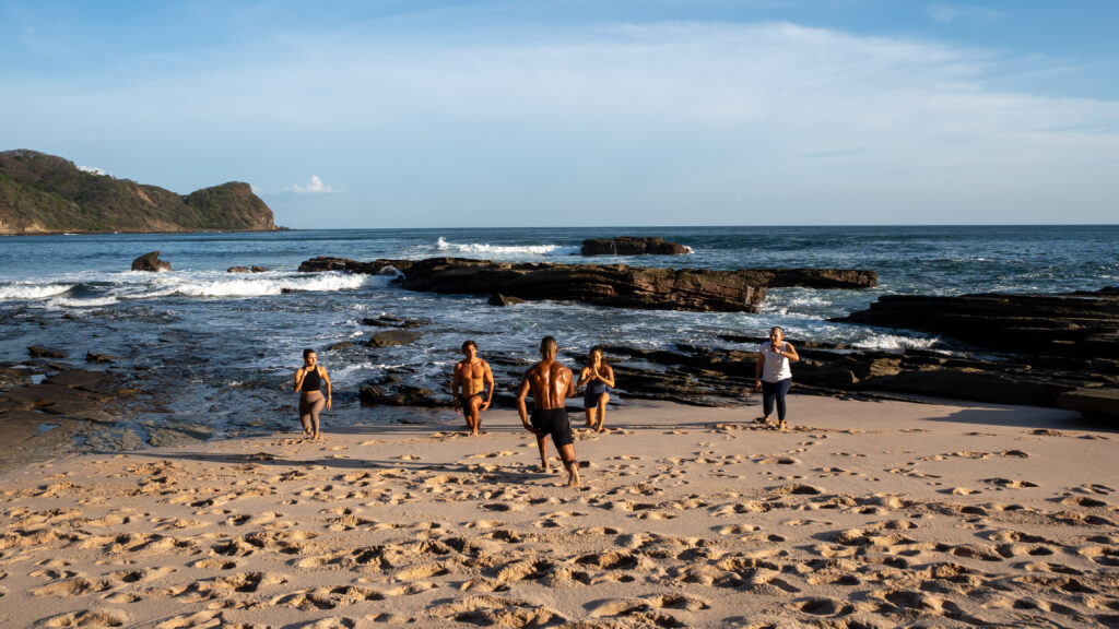 Five people stroll along a sandy beach near the shoreline, waves crashing on rocks and a hilly coastline behind them—a serene moment captured on their FitSouls wellness journey under the clear blue sky.