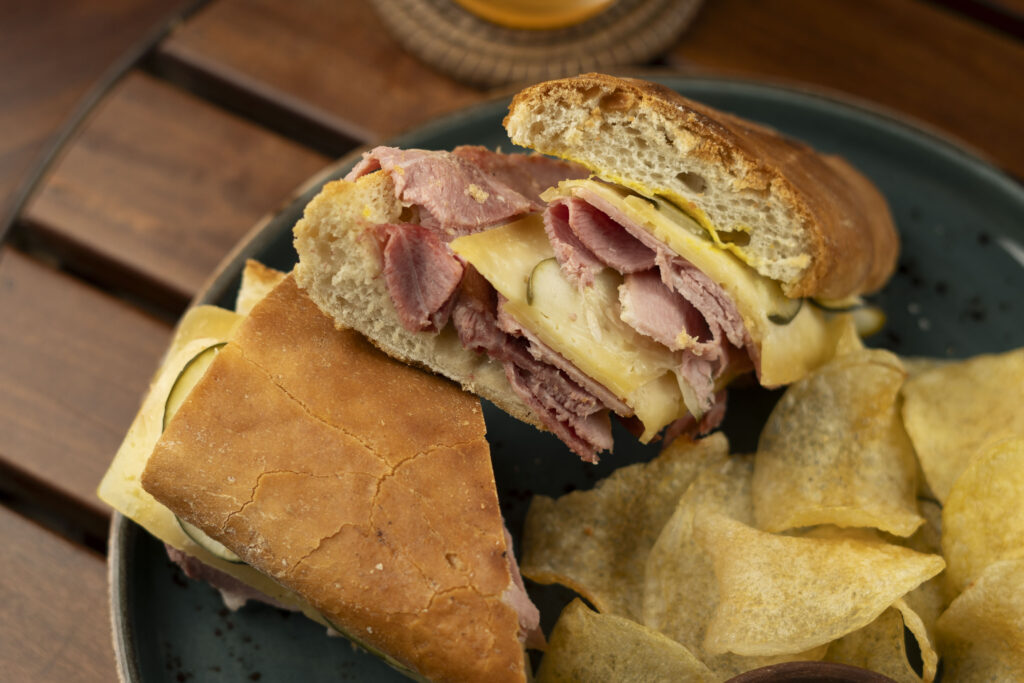 A close-up of a sandwich with sliced meat, cheese, and pickles on crusty bread, served on a plate with potato chips—perfect for a delicious Sunday Brunch.