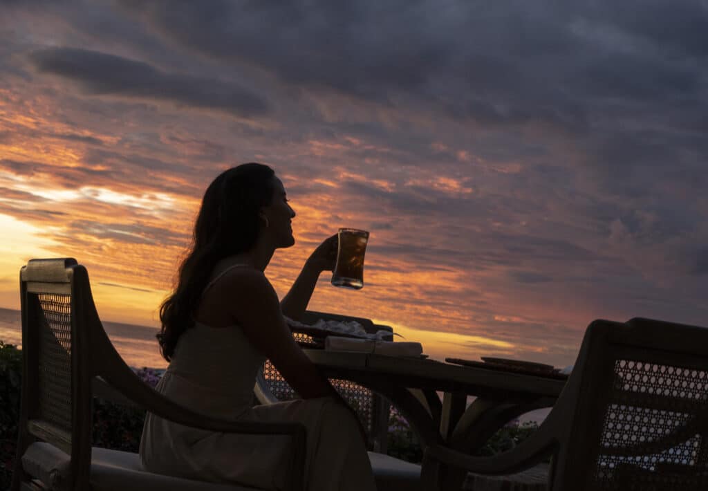A woman in a dress sits at an outdoor table on the Nicaraguan coast, holding a drink and gazing at a dramatic sunset sky with orange and purple clouds. The scene, enhanced by subtle Mexican flavors, is peaceful and relaxing.