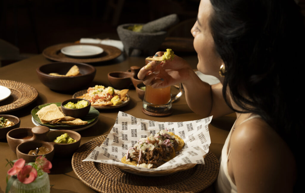 A woman sits at a table enjoying bold Mexican flavors, holding a taco. Various dishes such as chips, guacamole, and salads are spread out on woven placemats, with a drink and decorative flower also visible.