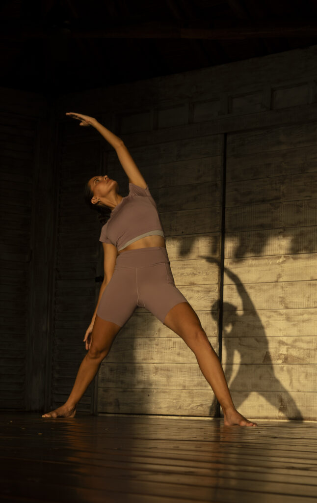 A woman in mauve workout clothes performs a yoga stretch, reaching one arm overhead while standing in a wide-legged pose against a wooden wall lit by warm sunlight, embodying her wellness journey with FitSouls.