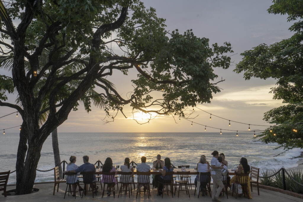 A group of people sit at a long table under string lights and large trees by the sea, facing the ocean and watching the sunset. The scene is tranquil and social, with a scenic view of the beach and water.