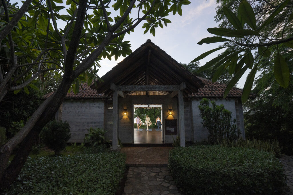 A rustic building with a tiled roof and lanterns at the entrance stands among lush greenery. Through the open doorway, people are visible practicing yoga or dancing inside.