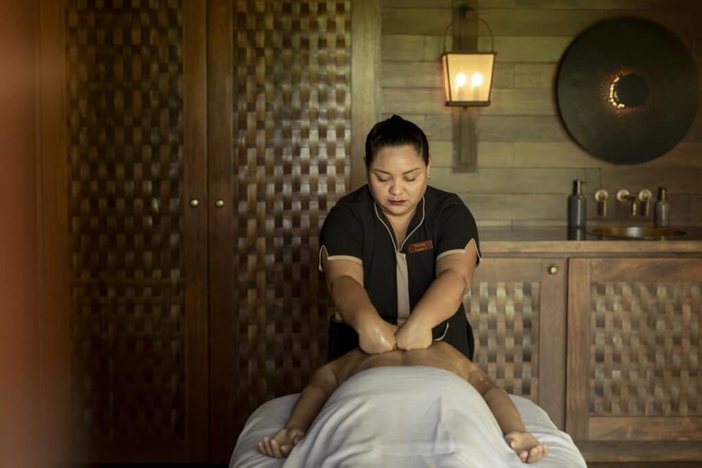 A massage therapist in a dark uniform gives a back massage to a person lying face-down on a table in a warmly lit, wooden spa room.