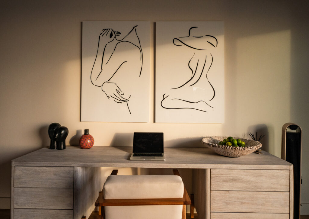 A modern desk setup with a closed laptop, decorative items, and a bowl of green apples. Above the desk are two minimalist line art prints depicting abstract human figures. Warm sunlight fills the room.