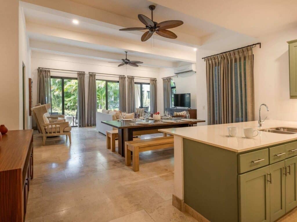 Open-concept living space with a kitchen island perfect for family pizza nights, a dining table, and a comfortable seating area. Large windows with views of palm trees, ceiling fans, neutral tones, and natural light create a bright, inviting atmosphere ideal for family cooking or hosting your own pizza-making class.