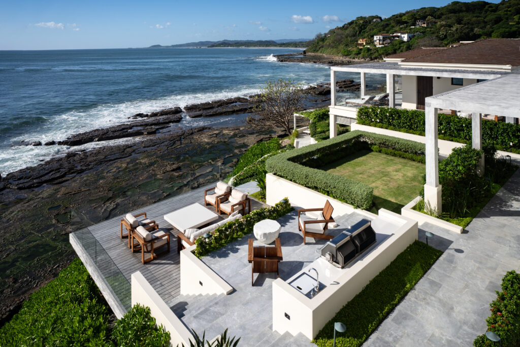 A modern seaside patio with outdoor seating, a grill, and a dining area overlooks rocky shores and the ocean, surrounded by lush greenery and a manicured lawn under a partly cloudy sky.