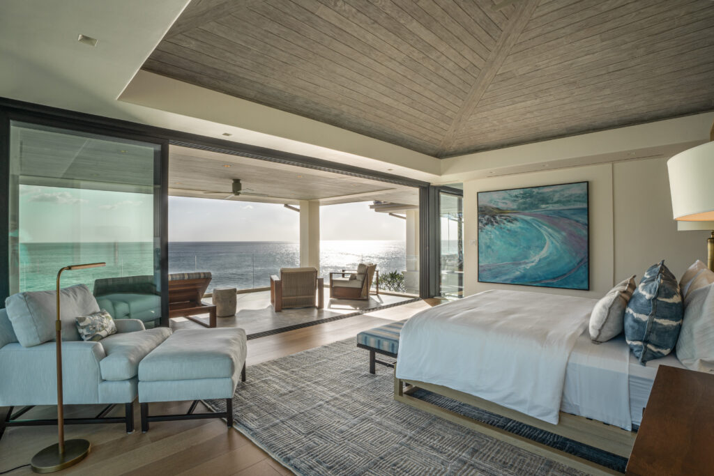 A luxurious bedroom with a large bed, armchair, and modern decor overlooks the ocean through floor-to-ceiling glass doors leading to a furnished balcony at sunset. Light fills the room, highlighting a coastal painting on the wall.