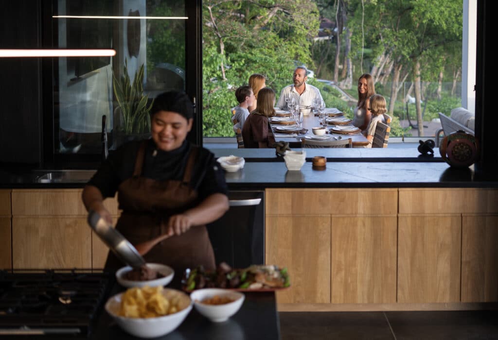 A chef prepares food in a modern kitchen while a family of six sits at an outdoor dining table, smiling and talking surrounded by greenery.