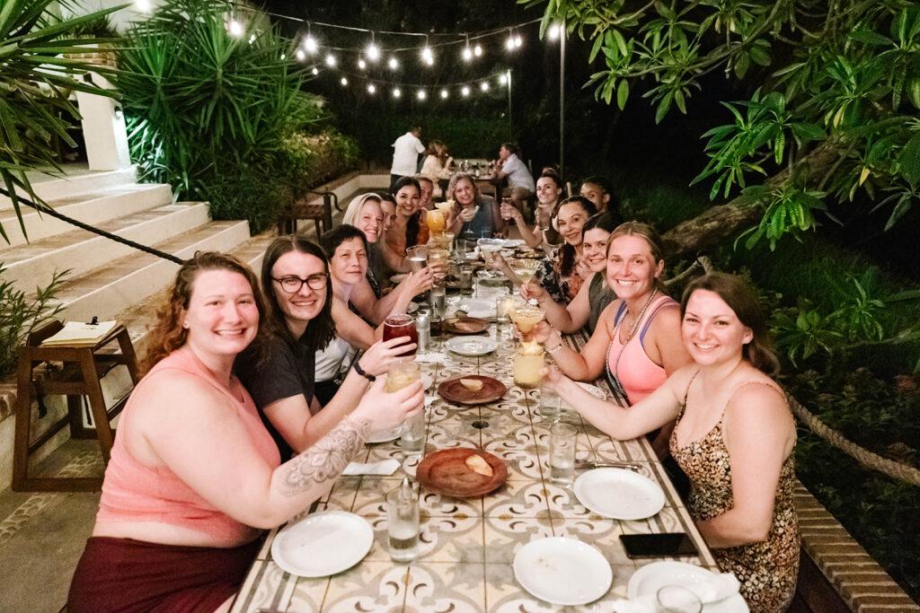 A group at a wellness retreat sits at a long, outdoor table under string lights at night, smiling and raising drinks together. The lush greenery and festive ambiance create a relaxed, joyful atmosphere.