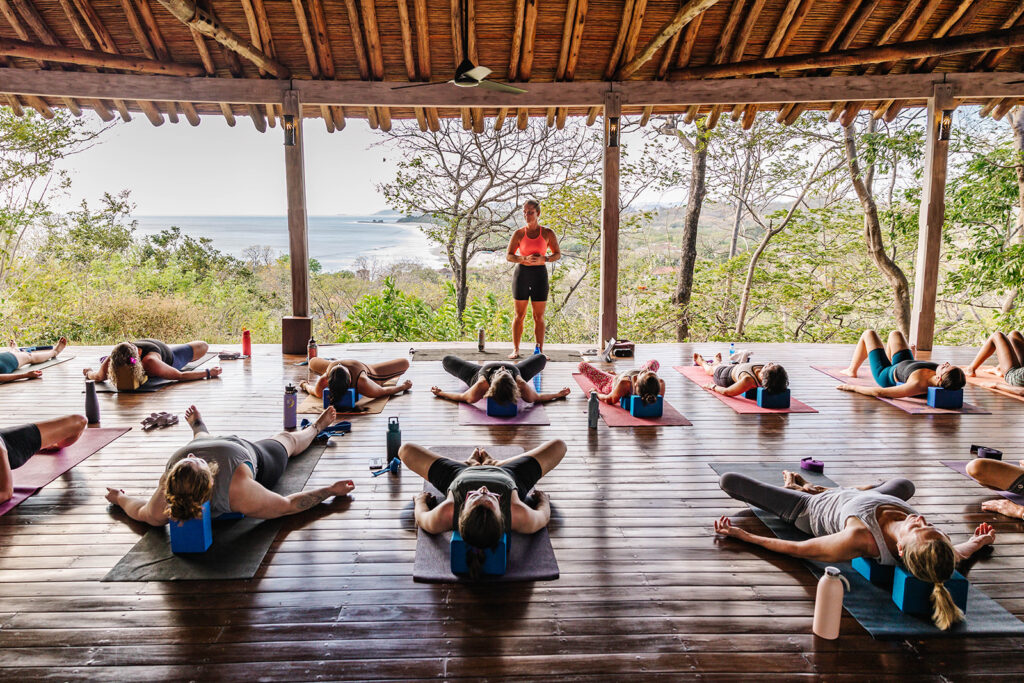 A group of people practice yoga on mats in a wooden pavilion overlooking nature and the ocean during a wellness retreat. A yoga instructor stands at the front, while participants lie on their backs in a relaxed pose, using yoga blocks.