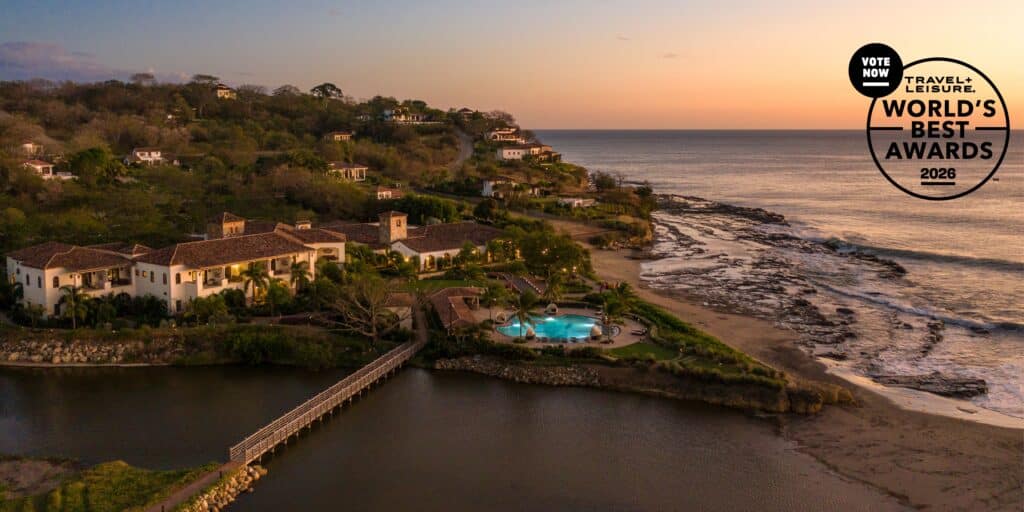 Aerial view of a coastal resort at sunset, with terracotta-roofed villas, a pool, lush greenery, and a wooden bridge over water. The ocean and rocky shoreline are visible, along with a Worlds Best Awards 2026 badge.