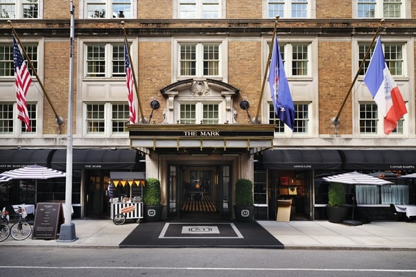 The entrance of The Mark Hotel, a historic brick building with flags, striped awnings, and black-and-gold doors flanked by trimmed bushes on a city sidewalk, is an icon for luxury travel and an anticipated hotspot in 2026 travel trends.