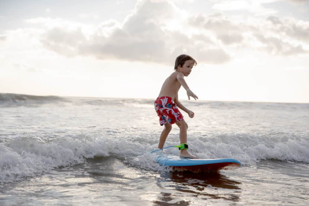A young child wearing red patterned swim shorts surfs a small wave on a blue surfboard in shallow ocean water, with a cloudy sky and gentle sunlight in the background.