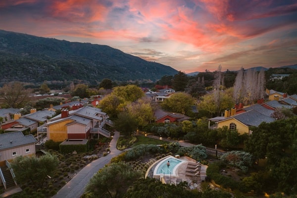 Aerial view of a residential area with colorful houses, lush trees, and a circular pool in the foreground—an inviting scene that reflects 2026 luxury travel trends against hills and a dramatic, colorful sunset sky.