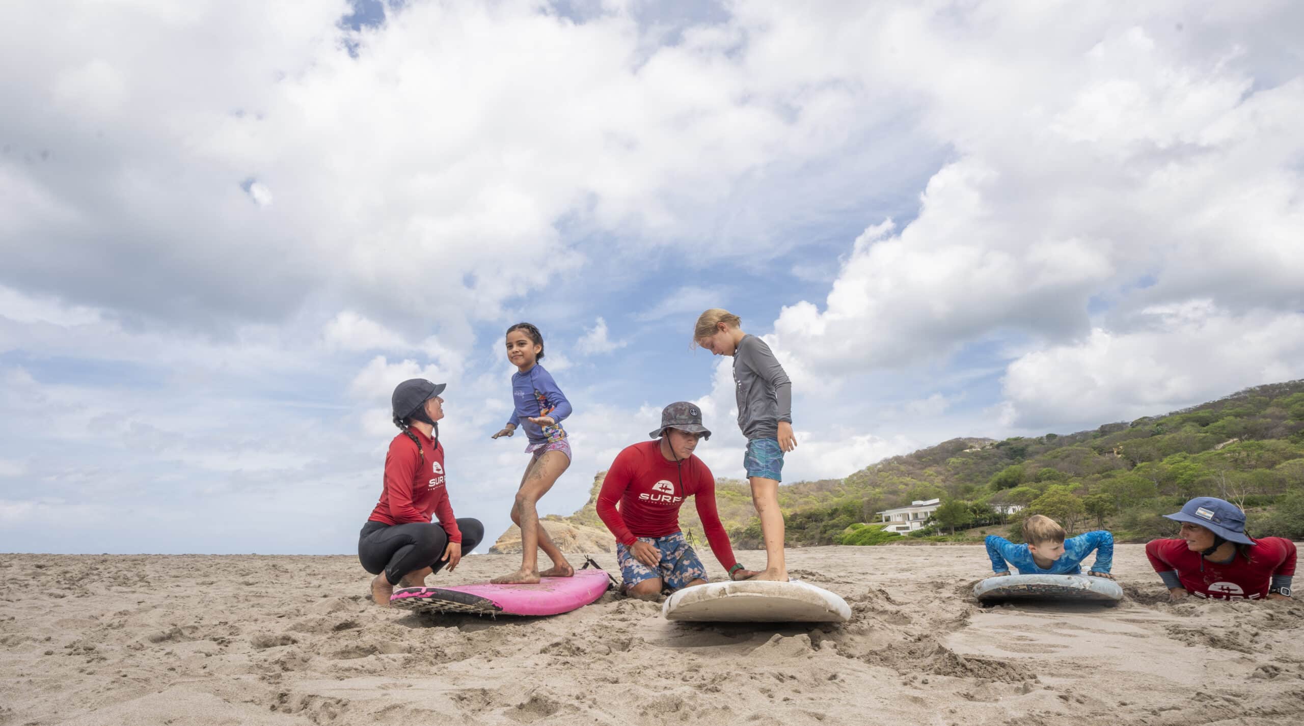 Four children and two adults practice surfing on the beach, standing or lying on boards in the sand under a cloudy sky, with green hills and a house visible in the background.