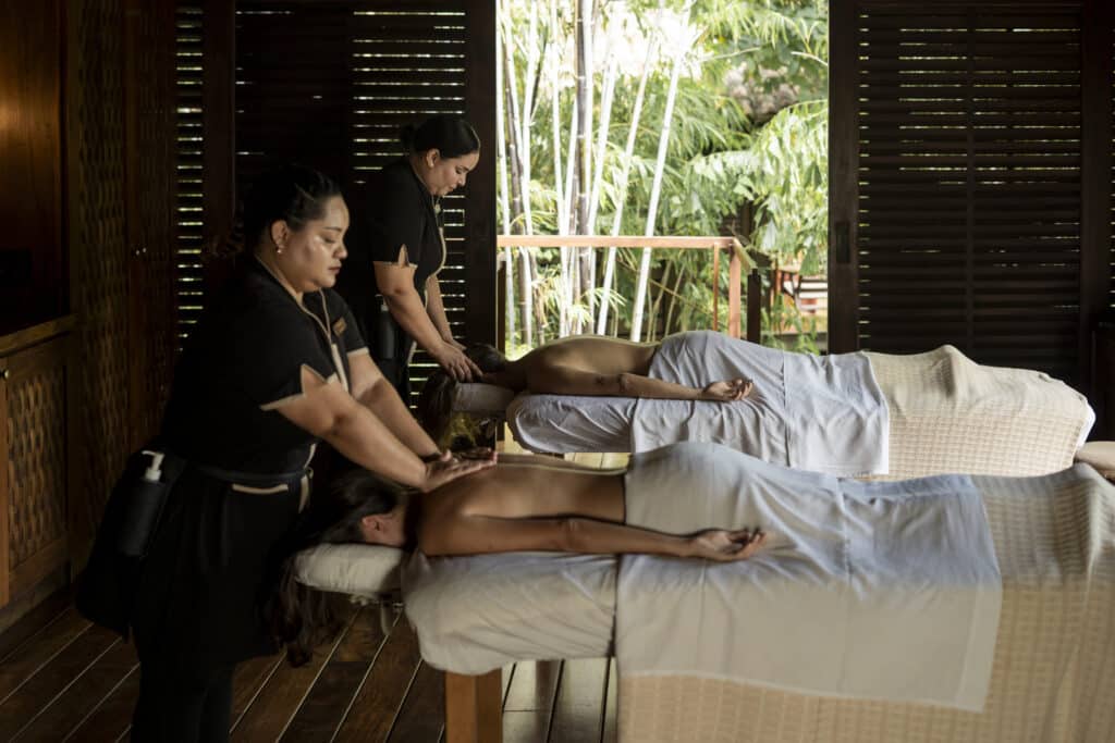 Two people receive massages on side-by-side tables in a serene, wooden spa room with large windows revealing lush greenery outside. Two massage therapists work on each person, creating a relaxing atmosphere.