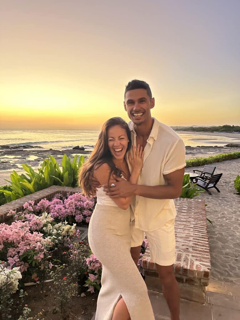 A smiling couple embraces in front of a beach at sunset, surrounded by pink flowers and greenery. The woman shows off an engagement ring as the International Souls bask in paradise, both wearing light-colored summer outfits.