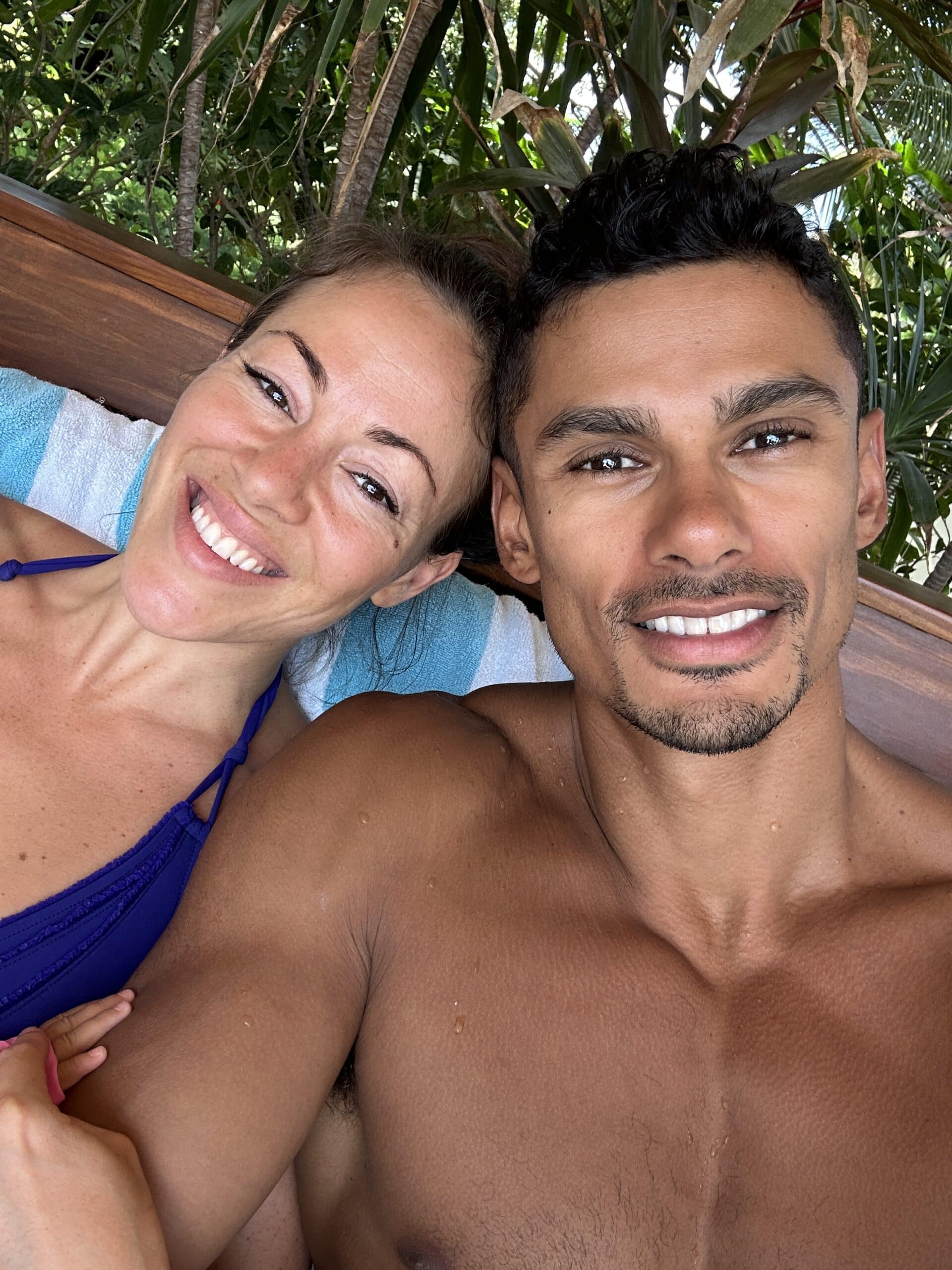 A smiling woman and man, truly International Souls, take a close-up selfie together outdoors, reclining on a blue and white towel with green tropical plants in the background.