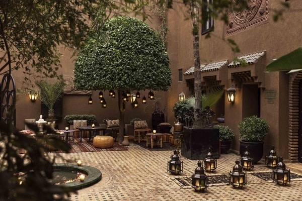 A Moroccan courtyard with patterned tile flooring, lanterns, potted plants, and a round tree. Cushioned seating surrounds a low table—an inviting scene reflecting 2026 luxury travel trends. Ornate lighting hangs from the tree, and the walls are warm terracotta.