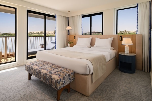 Modern hotel room with a large bed, white linens, a patterned bench at the foot, bedside tables with lamps, and large windows overlooking a river and trees. Light neutral tones reflect 2026 luxury travel trends for a calm, airy atmosphere.