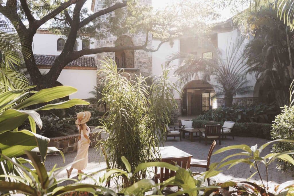 A woman in a sunhat and light dress enjoys a heartfelt homecoming as she walks through a lush, sunlit courtyard with tropical plants, wooden tables, and chairs, surrounded by white stone buildings and tall trees.