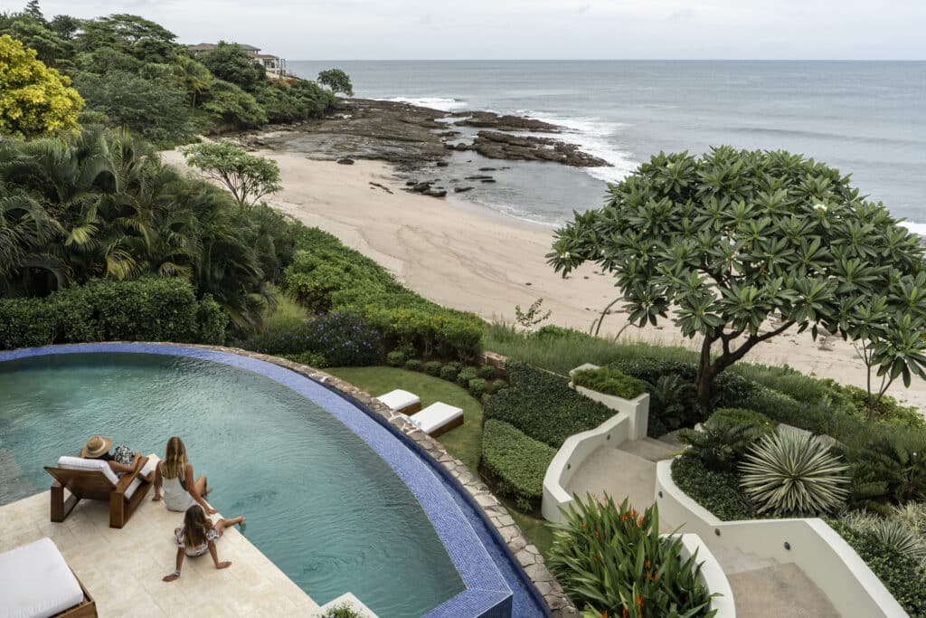 A woman and child relax by a curved pool overlooking a lush garden and a sandy beach with rocky shoreline, as calm ocean waves approach the shore—a heartfelt homecoming beneath a partly cloudy sky.